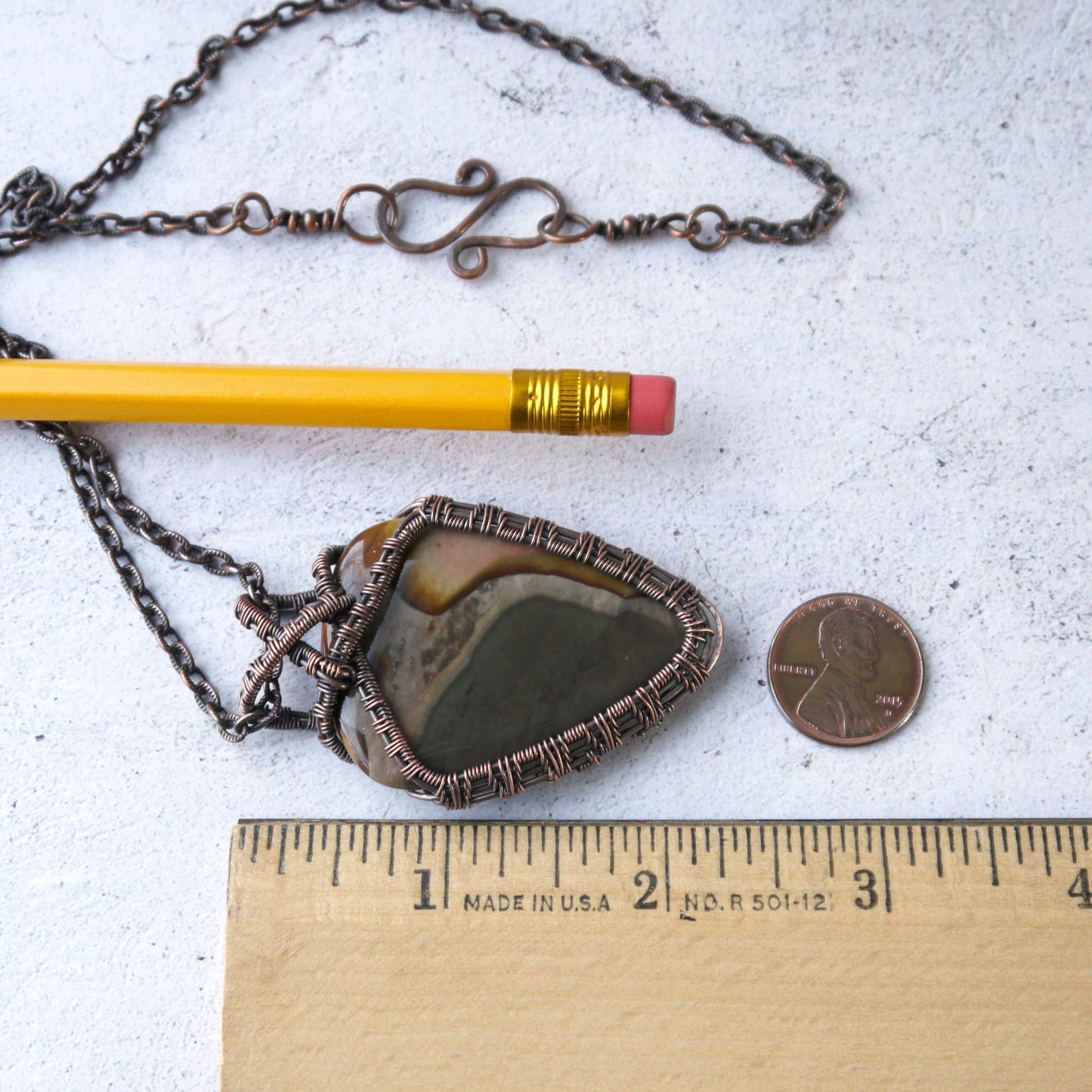 Necklace with a jasper copper wire wrapped handcrafted pendant next to a pencil and penny on a white surface