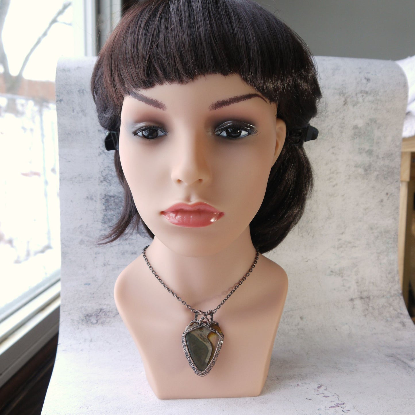 Mannequin head with a dark wig and impression jasper pendant copper necklace on a textured surface