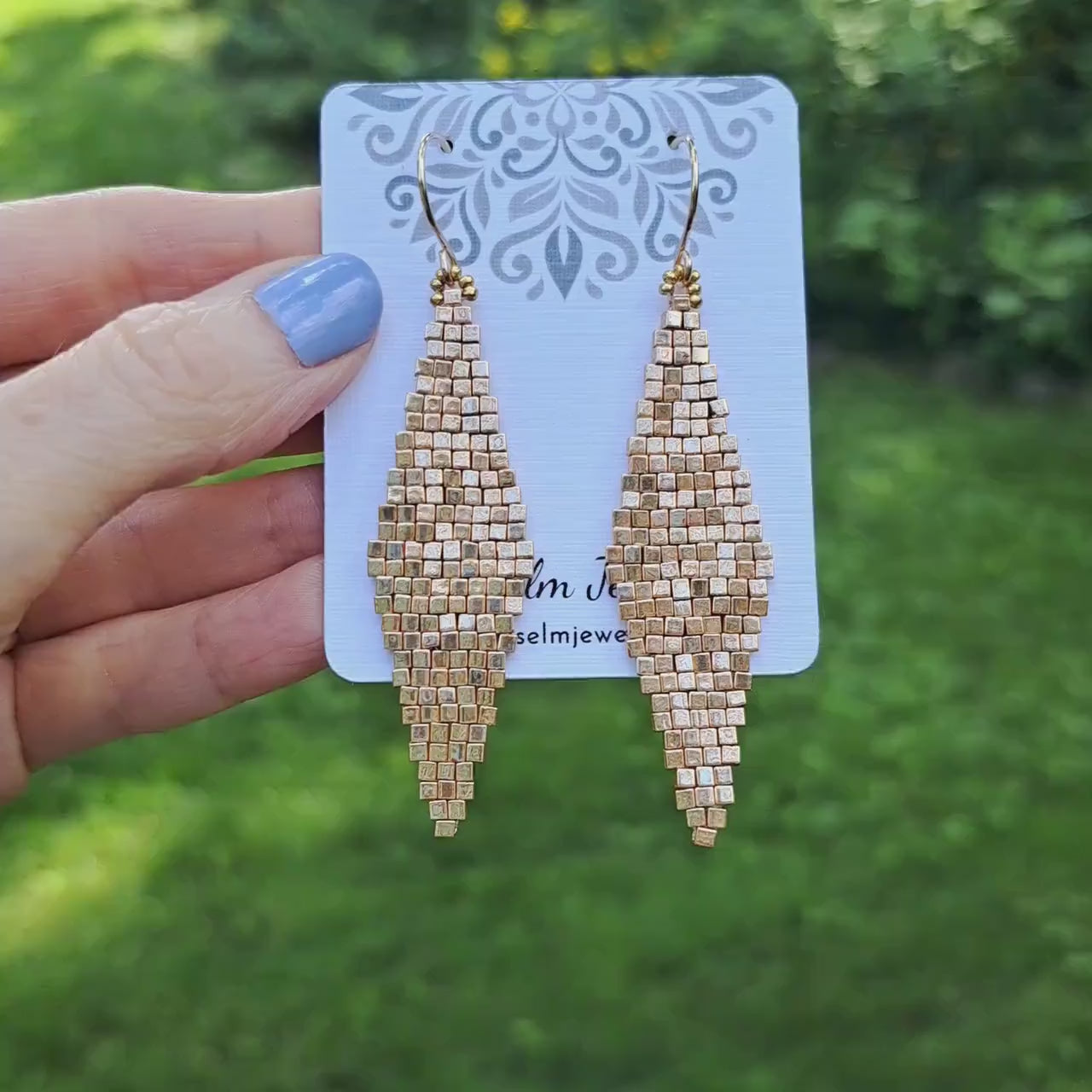 a pair of diamond shape woven earrings with cube shaped gold glass beads held in hand in outdoor setting