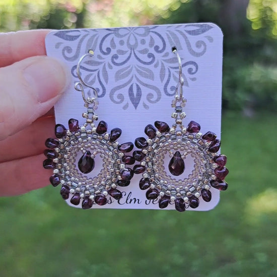 a pair of circular beadwoven garnet gemstone earrings on a Iris Elm Jewelry branded card held by hand