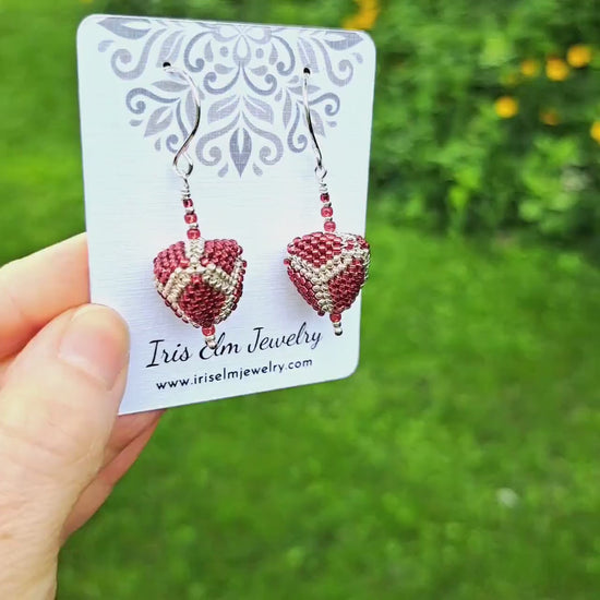 a pair of red and silver beadwork three dimensional triangle seed bead earrings on a 'Iris Elm Jewelry' branded card held in hand in outdoor setting