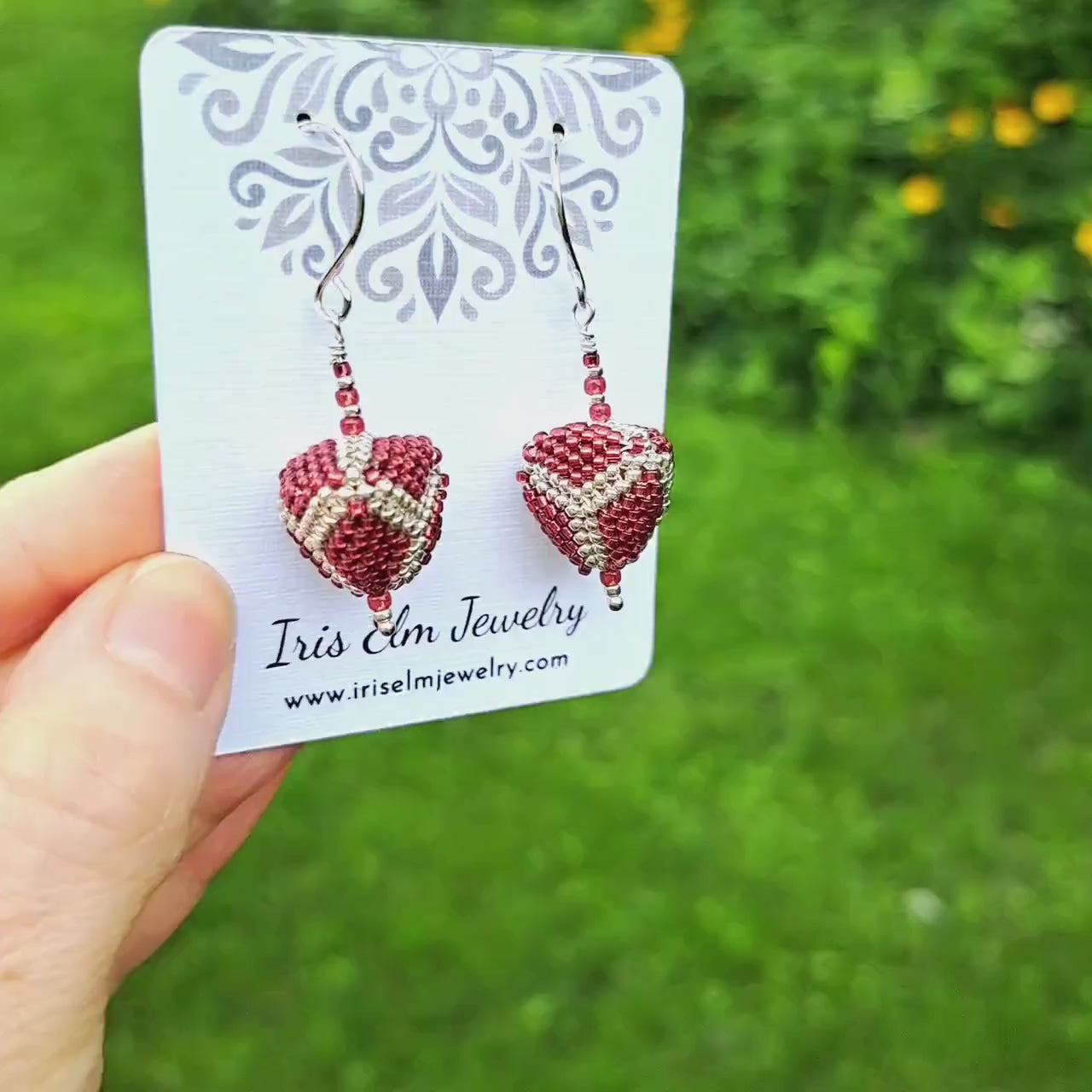 a pair of red and silver beadwork three dimensional triangle seed bead earrings on a 'Iris Elm Jewelry' branded card held in hand in outdoor setting