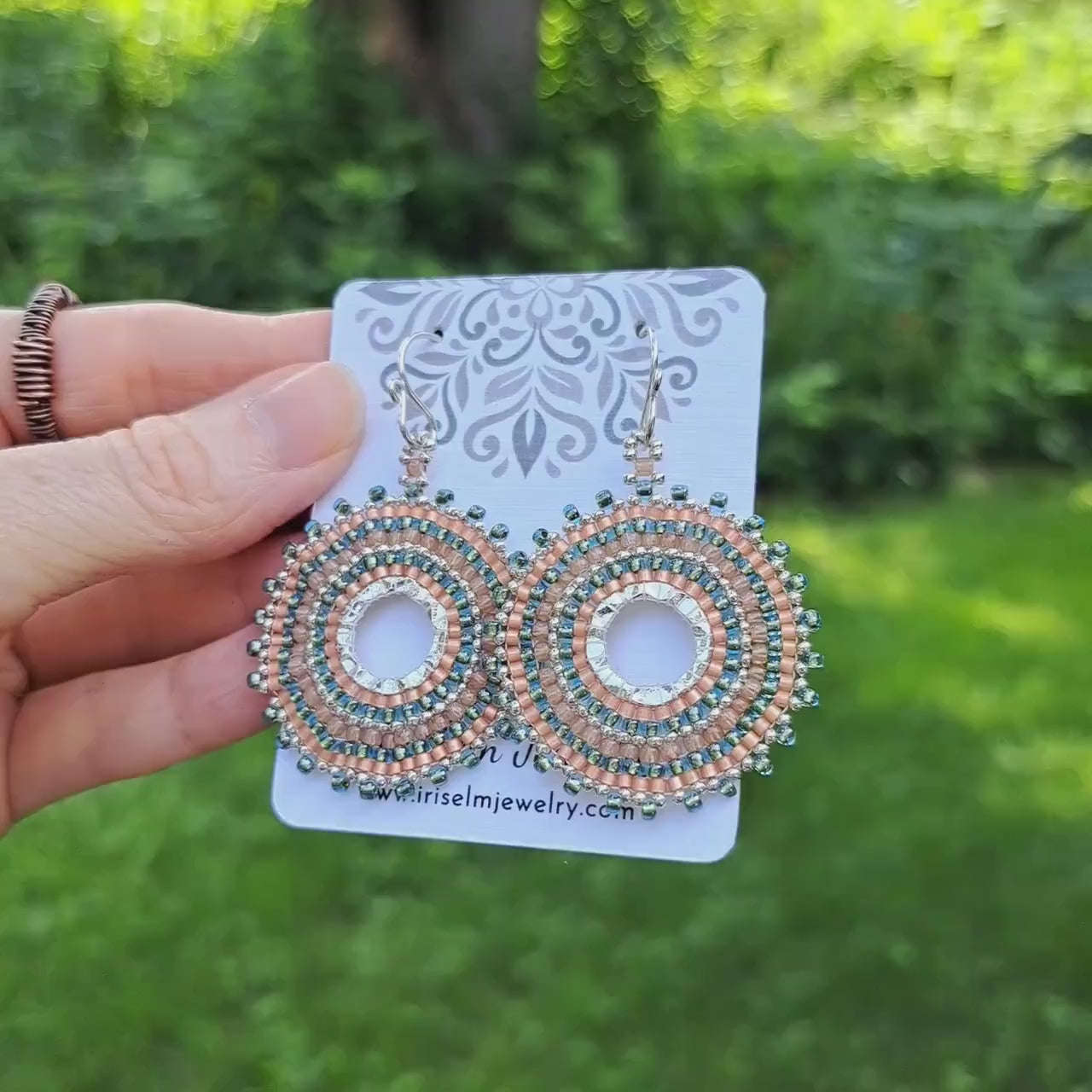 a pair of beadwoven aqua, peach, and silver seed bead hoop earrings on a 'Iris Elm Jewelry' card held by hand in outdoor setting