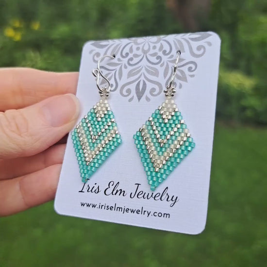 a pair of mint green, silver, and white chevron diamond shape seed bead earrings on a white 'Iris Elm Jewelry' branded card held by hand in outdoor setting