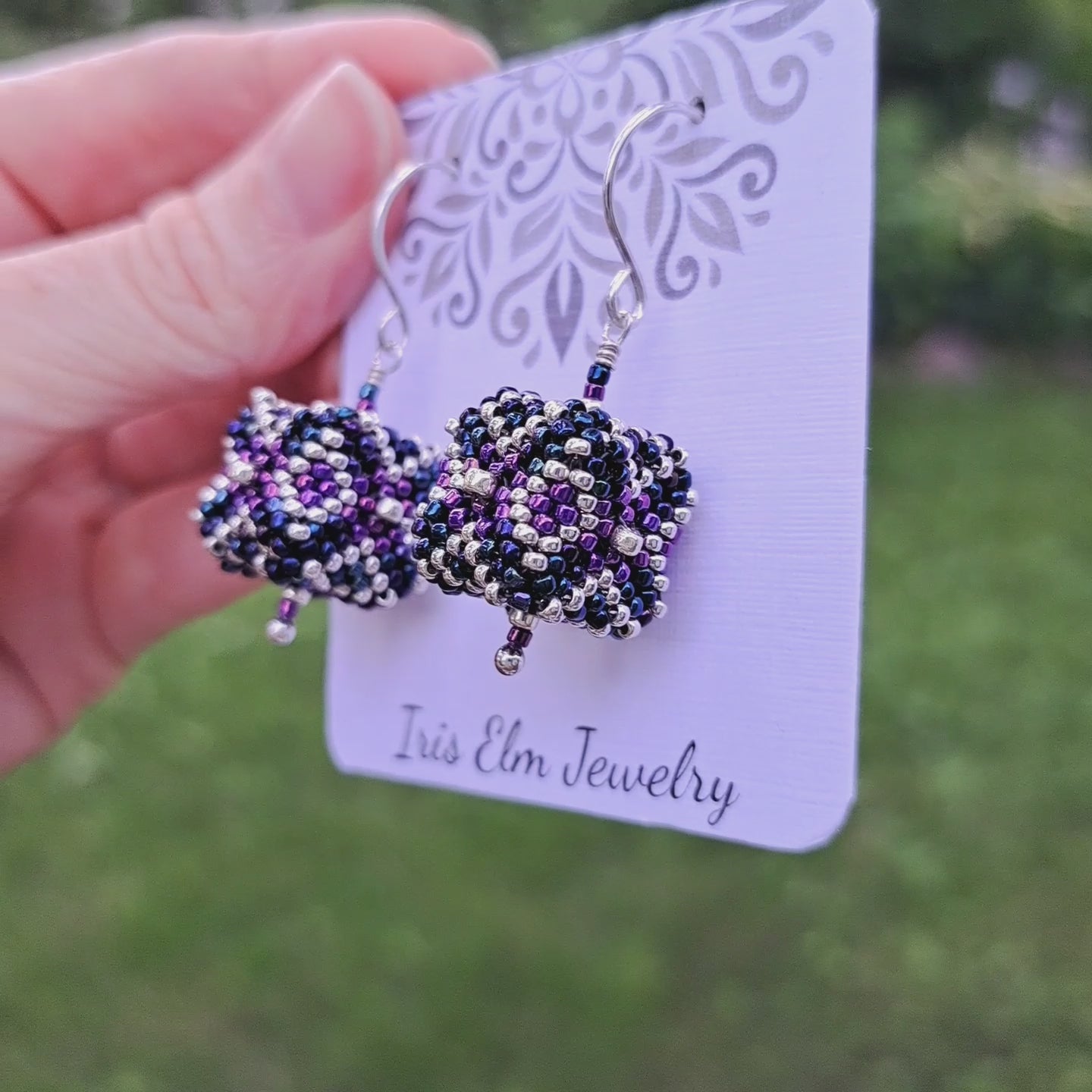 a pair of three-dimensional cube shaped purple, blue, and silver seed bead earrings on a 'Iris Elm Jewelry' branded card held in hand