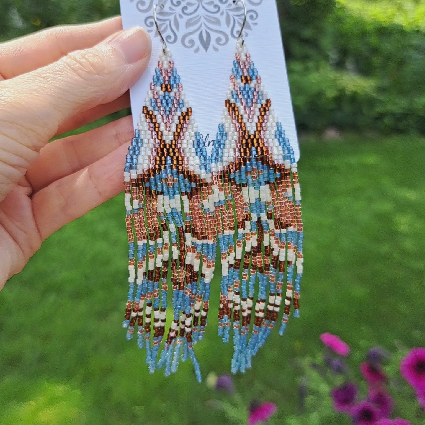 a pair of long seed bead blue, brown, white, and copper orange geometric fringe earrings on card held by hand in outdoor setting