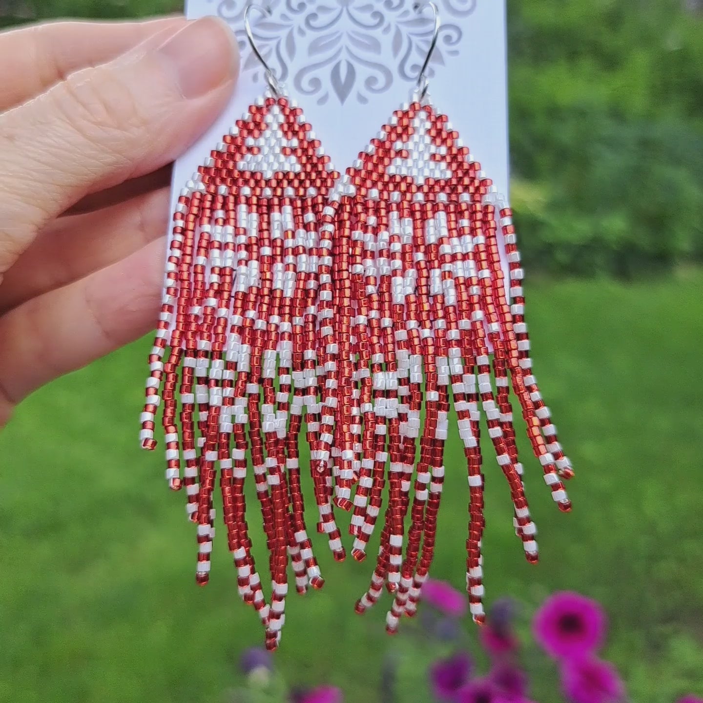 a pair of red and white snowflake patterned seed bead fringe earrings on card held in hand