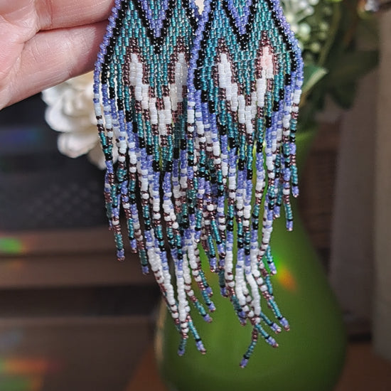 hand holding long purple, teal, black, and white seed bead heart fringe earrings