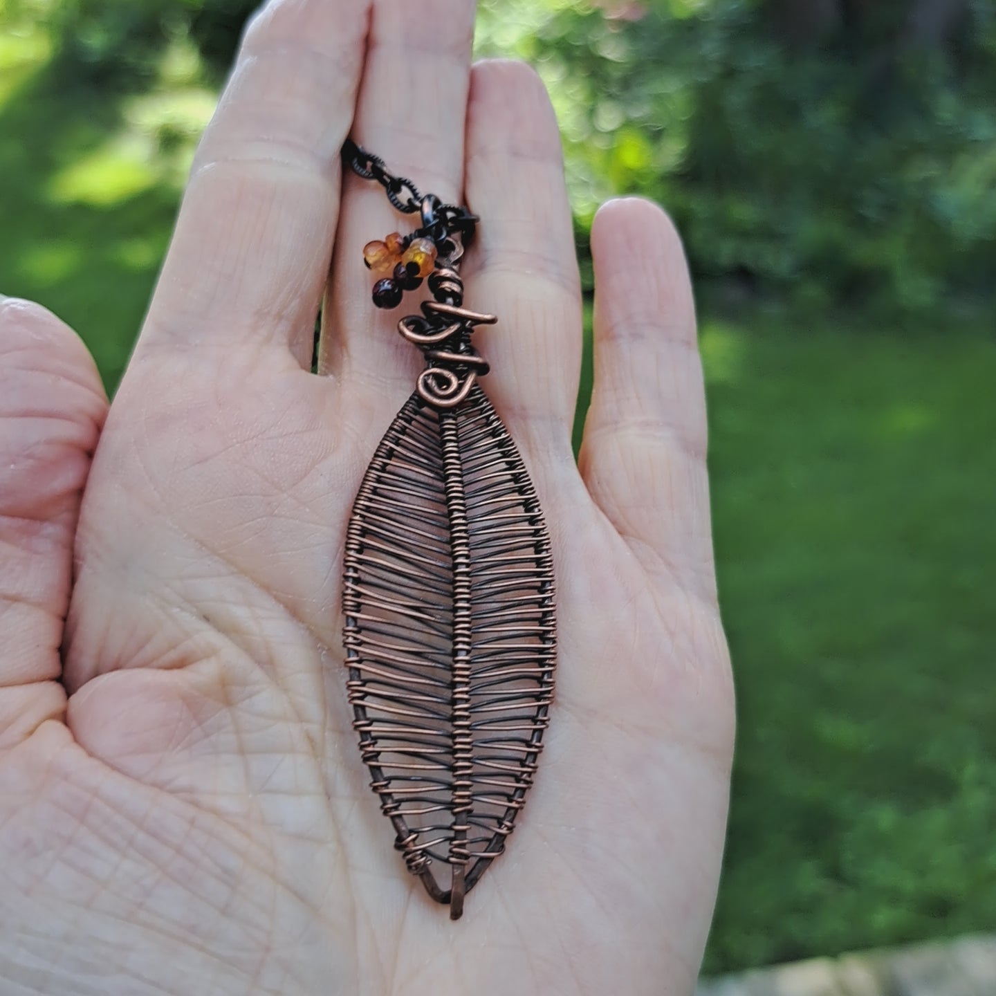 hand holding a copper wire wrapped leaf shaped pendant necklace with garnet and carnelian beads in outdoor setting
