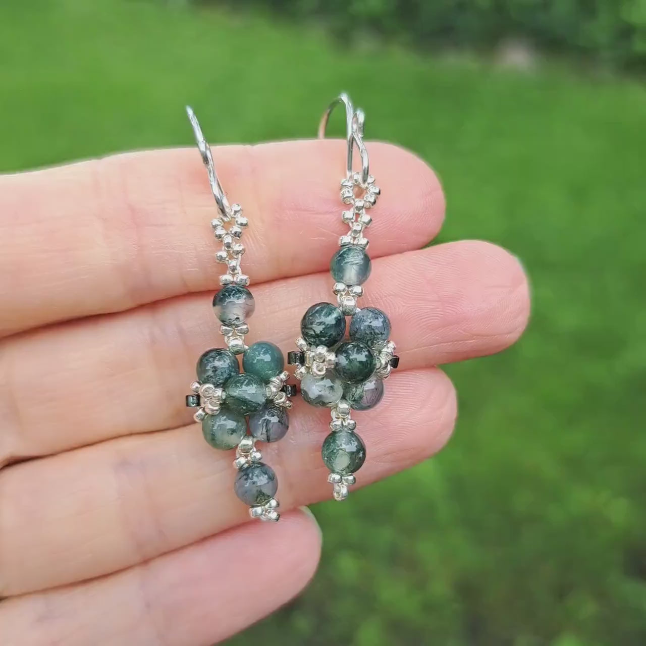 a pair of green moss agate beadwoven earrings held in hand in outdoor setting
