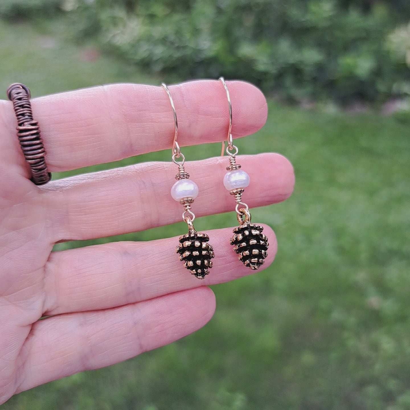 pair of earrings with gold toned pinecones and white freshwater pearls held in hand in outdoor setting