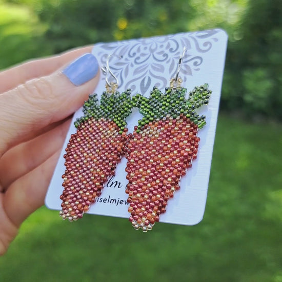 a pair of beadwoven red strawberry seed bead earrings on card held by hand in outdoor setting