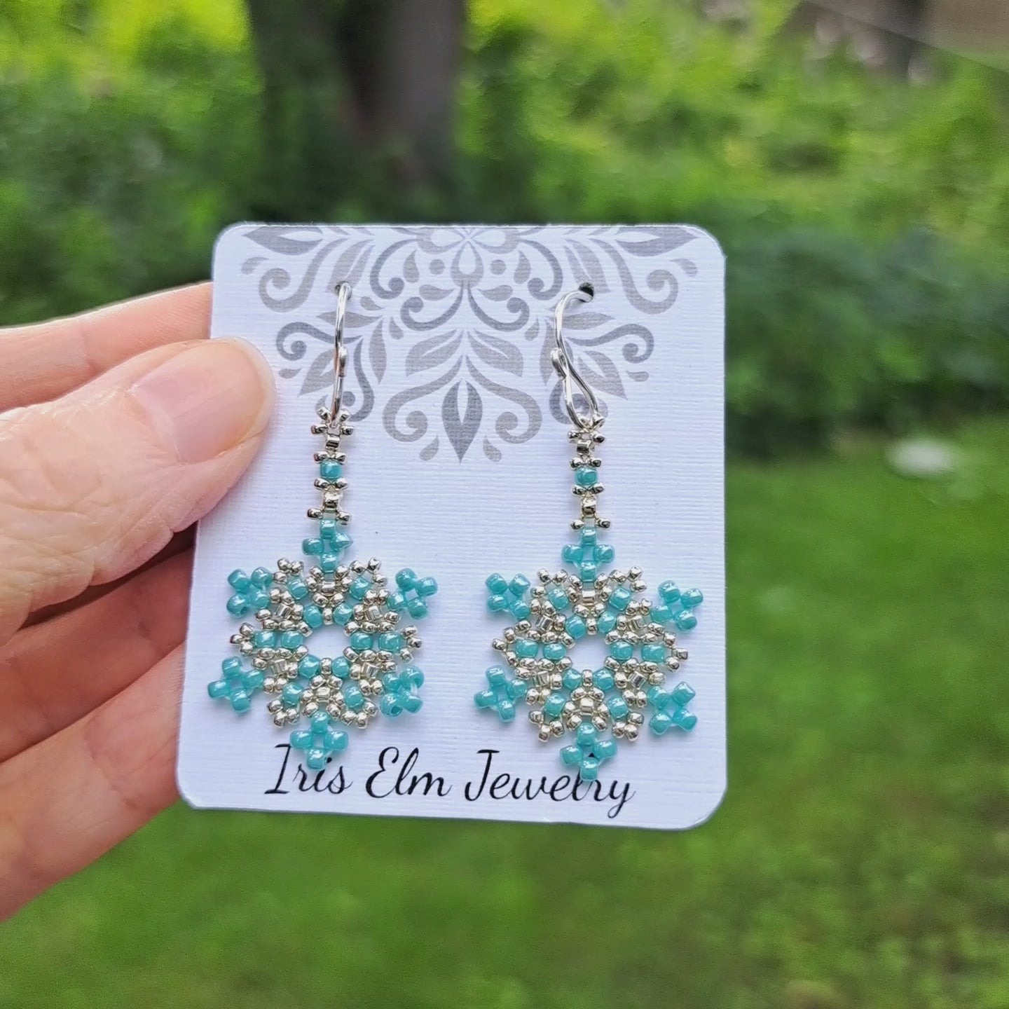 a pair of seed bead turquoise and silver snowflake earrings on Iris Elm Jewelry branded card held by hand