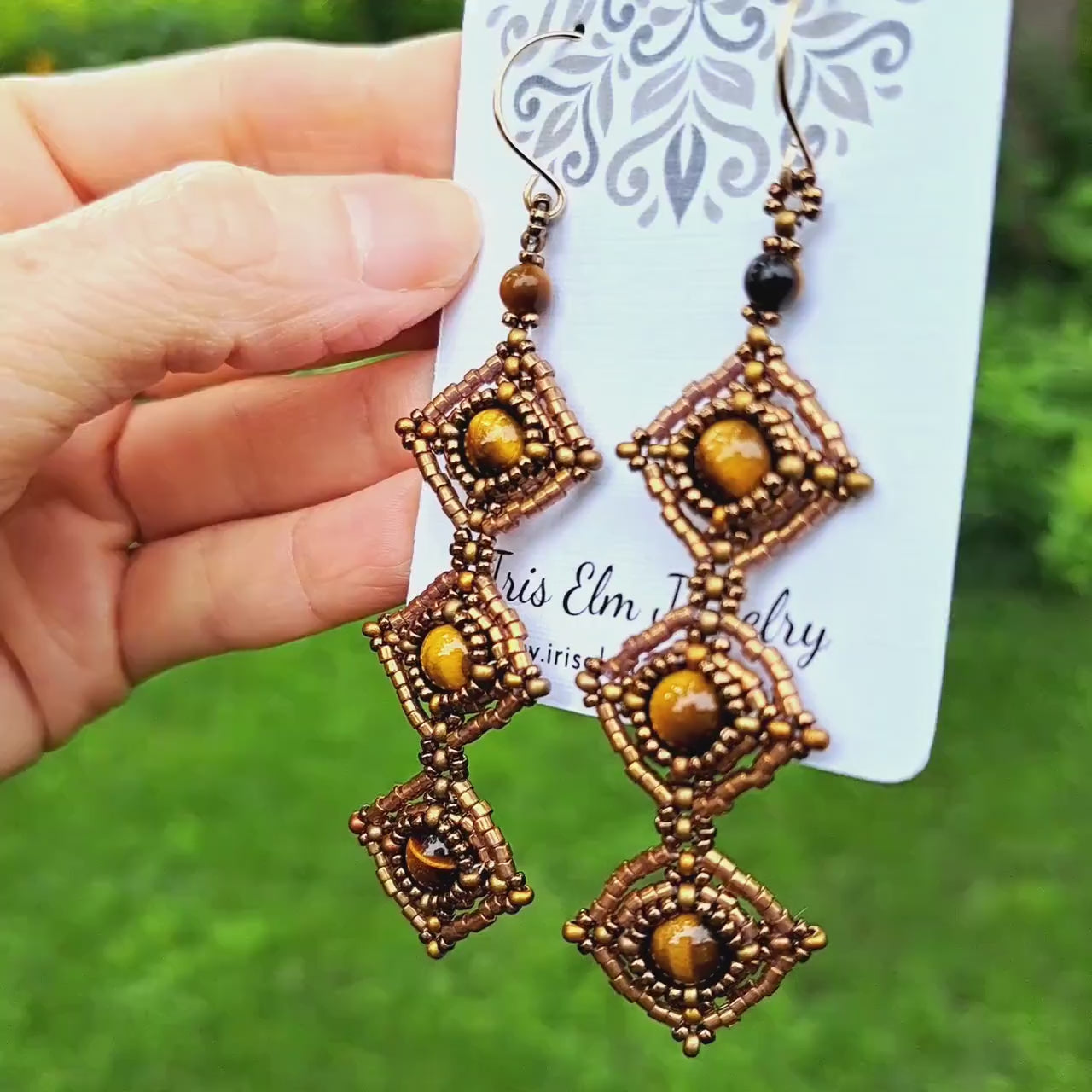 a pair of beadwork earrings woven with bronze seed beads and a trio of cascading tiger eye gemstones. Earrings are on card held in hand in outdoor greenery setting