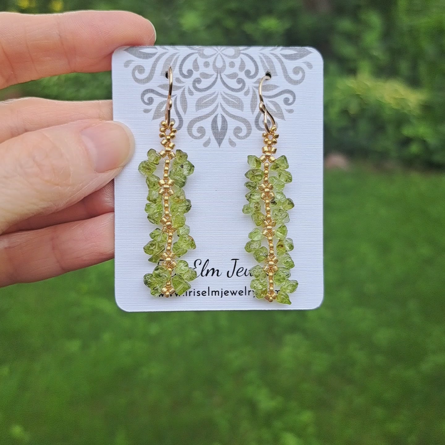 hand holding a Iris Elm Jewelry branded card with green peridot gemstone chip and gold glass seed bead beadwork earrings