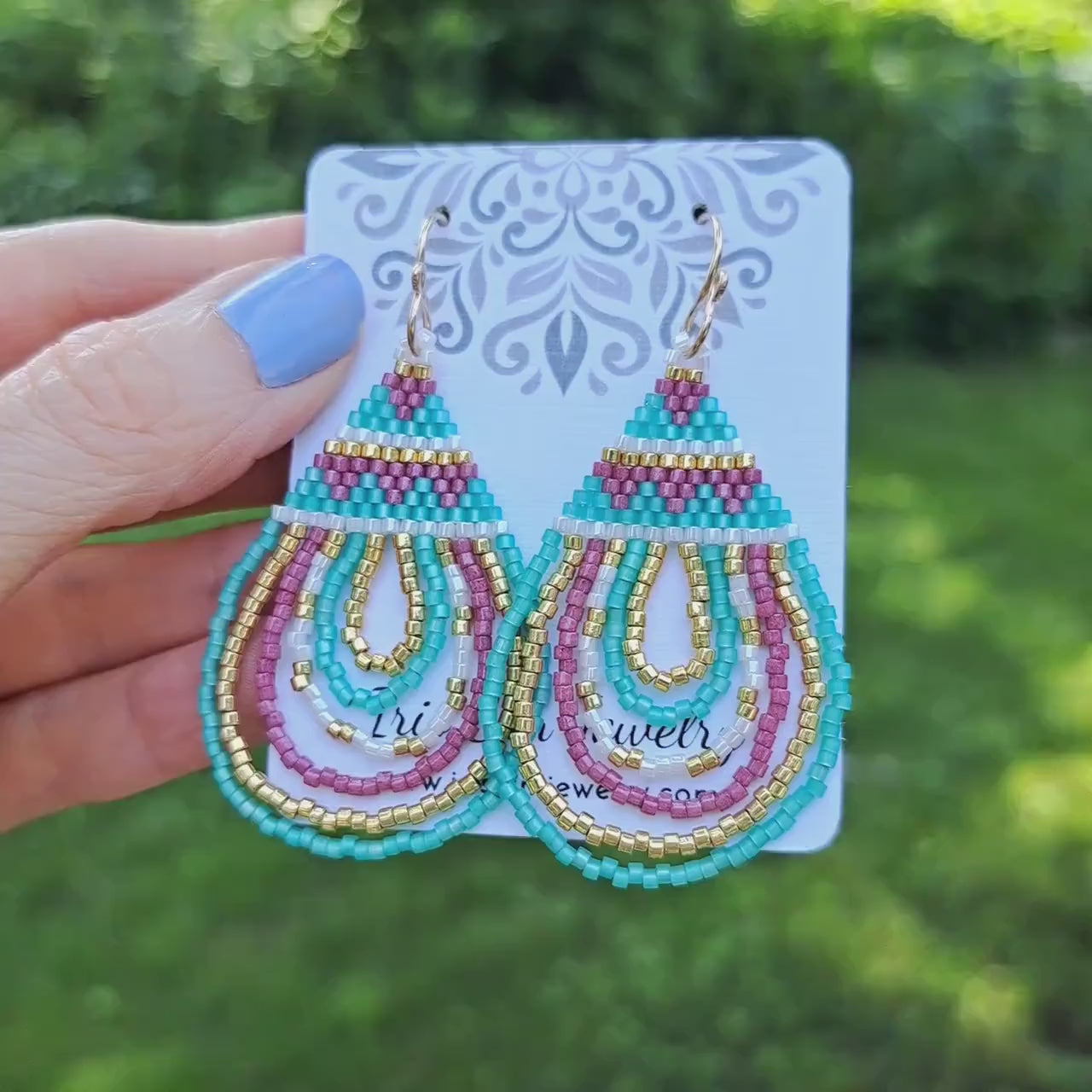 a pair of pink, mint green, and gold geometric teardrop seed bead earrings on a white card that is held by hand