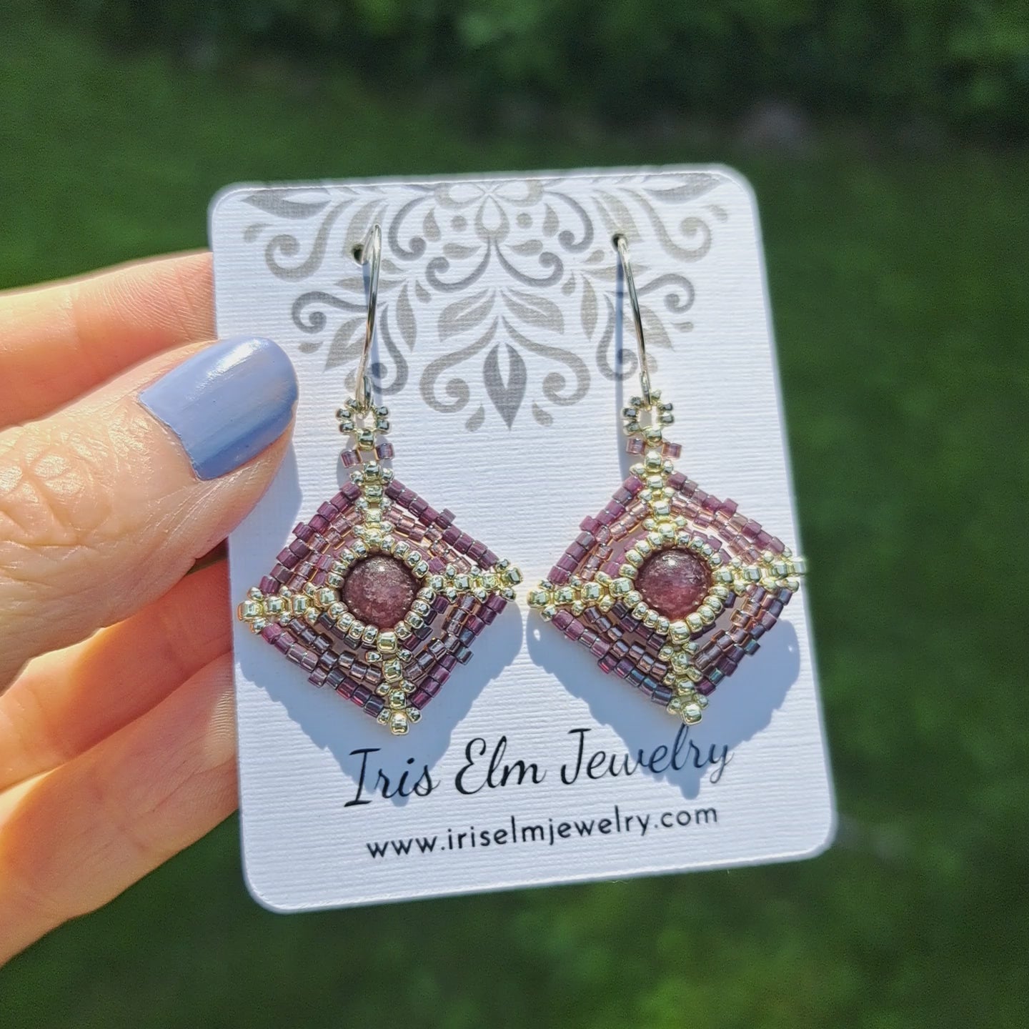 a pair of lepidolite beadwork earrings on a Iris Elm Jewelry branded card held by hand in outdoor setting