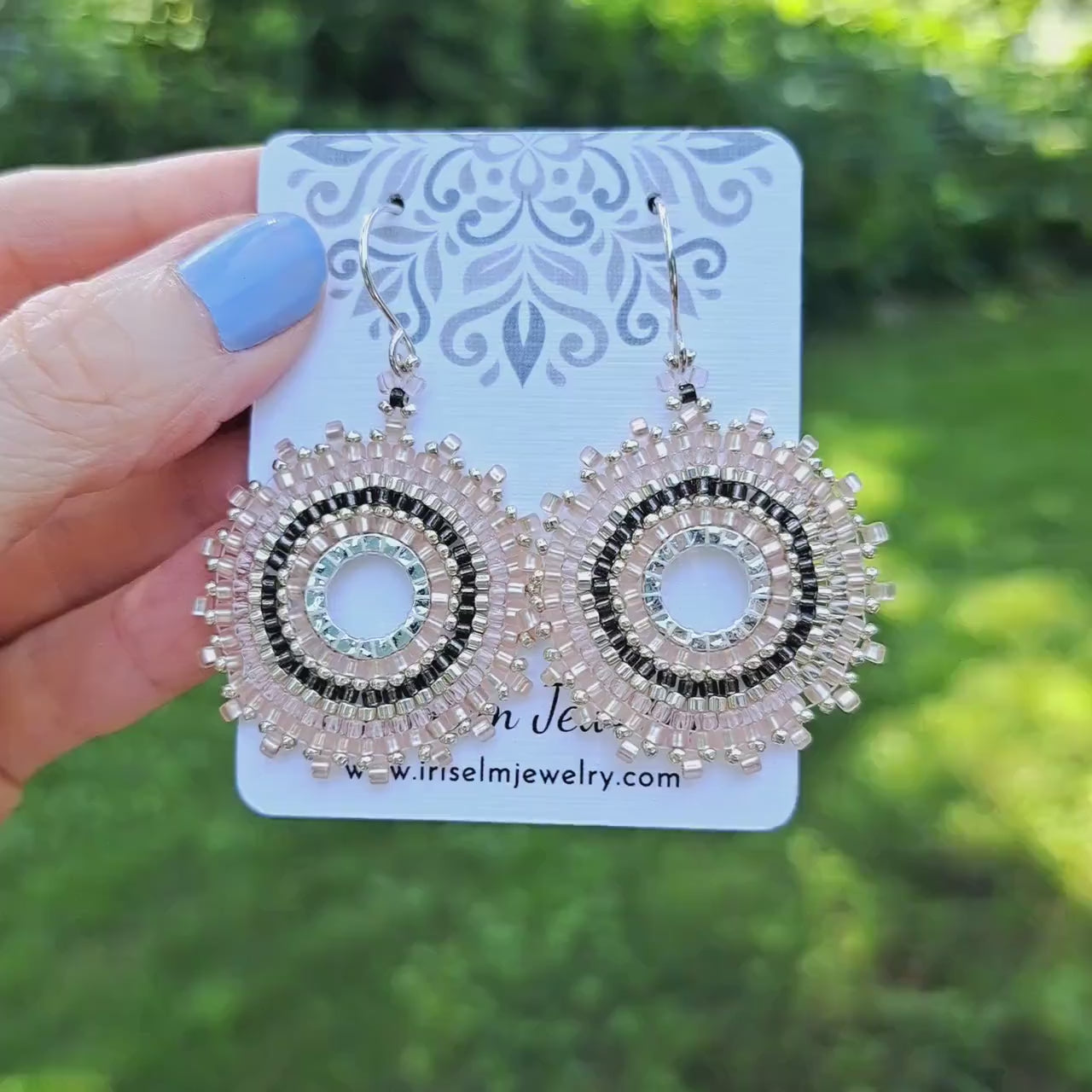 a pair of pale pink and dark grey glass seed bead woven hoop earrings on a 'Iris Elm Jewelry' branded card held by hand in outdoor setting