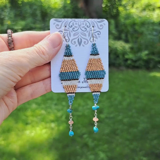 a pair of blue, bronze, white, and copper striped seed bead kite earrings with turquoise and sunstone dangles on card held by hand in outdoor setting