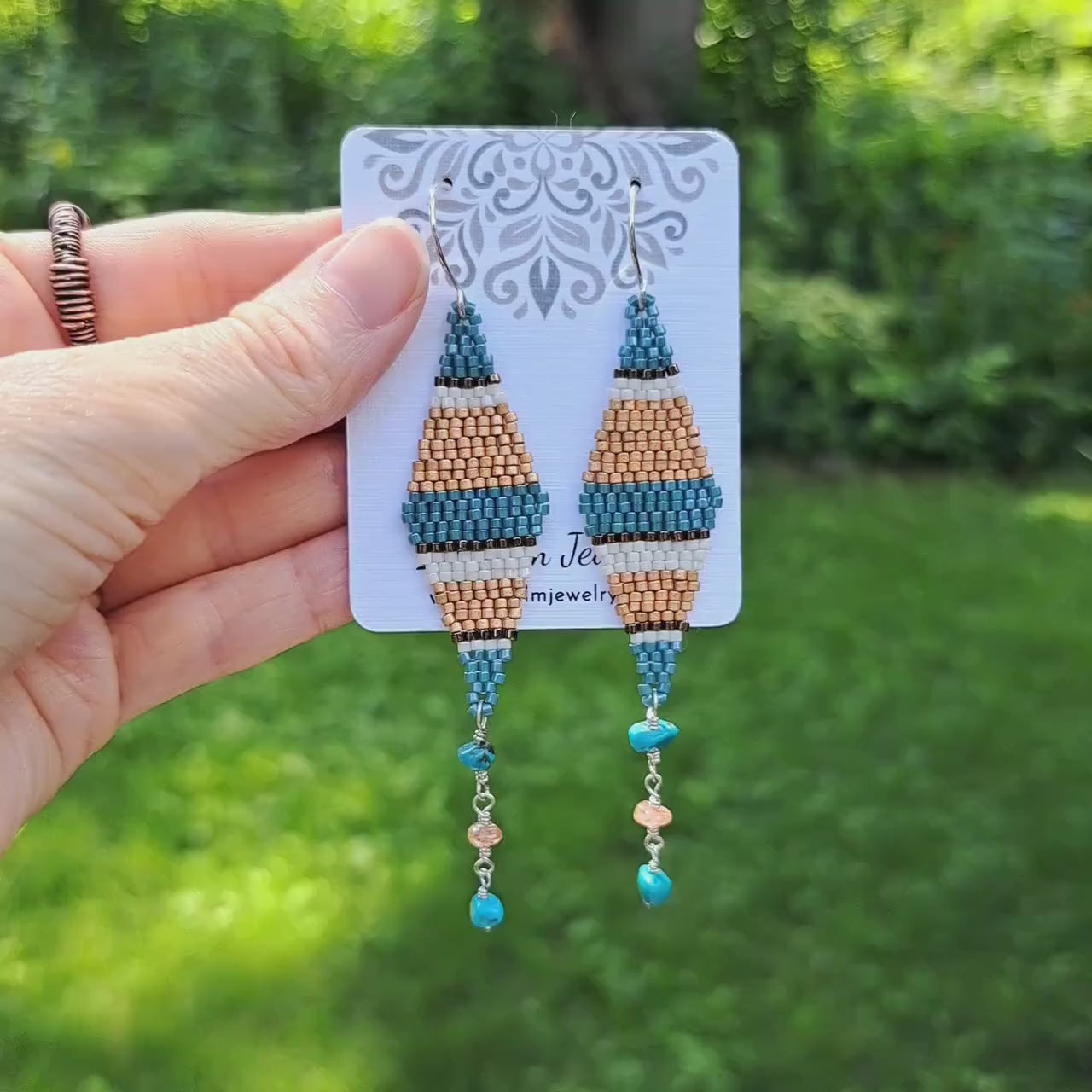 a pair of blue, bronze, white, and copper striped seed bead kite earrings with turquoise and sunstone dangles on card held by hand in outdoor setting