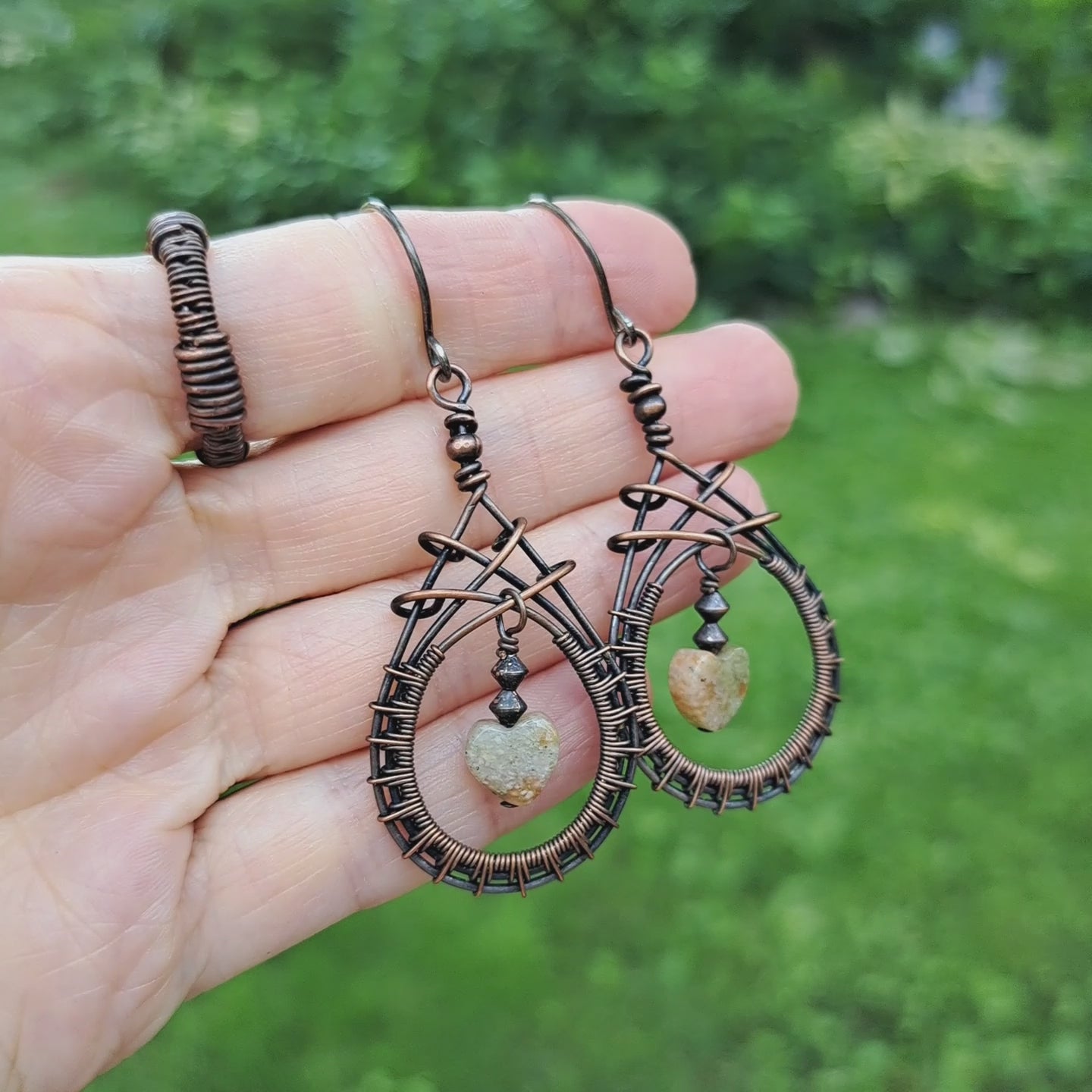 a pair of copper wire woven teardrop earrings with peach green autumn jasper heart shaped stones in center. earrings are held in hand in outdoor setting