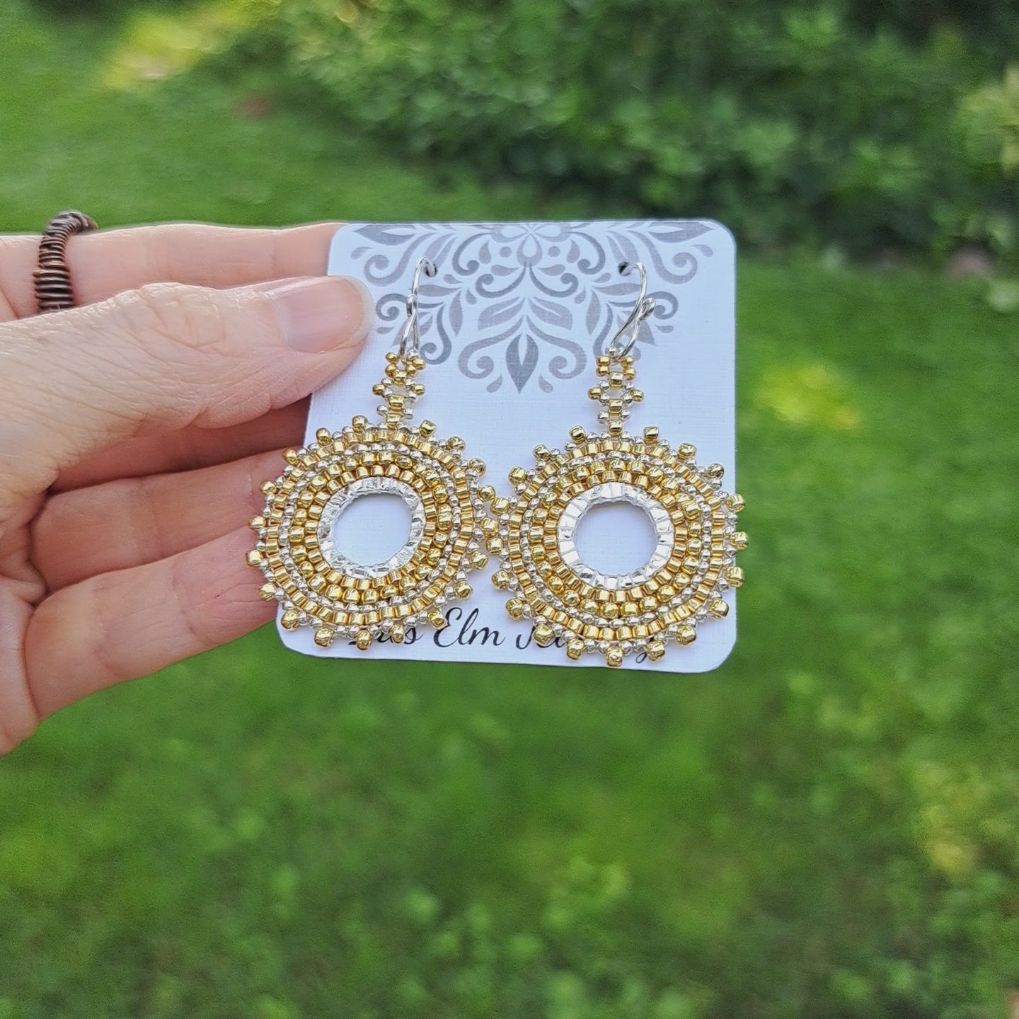 pair of gold and silver seed bead woven hoop earrings shown on 'Iris Elm Jewelry' card held by hand in outdoor setting