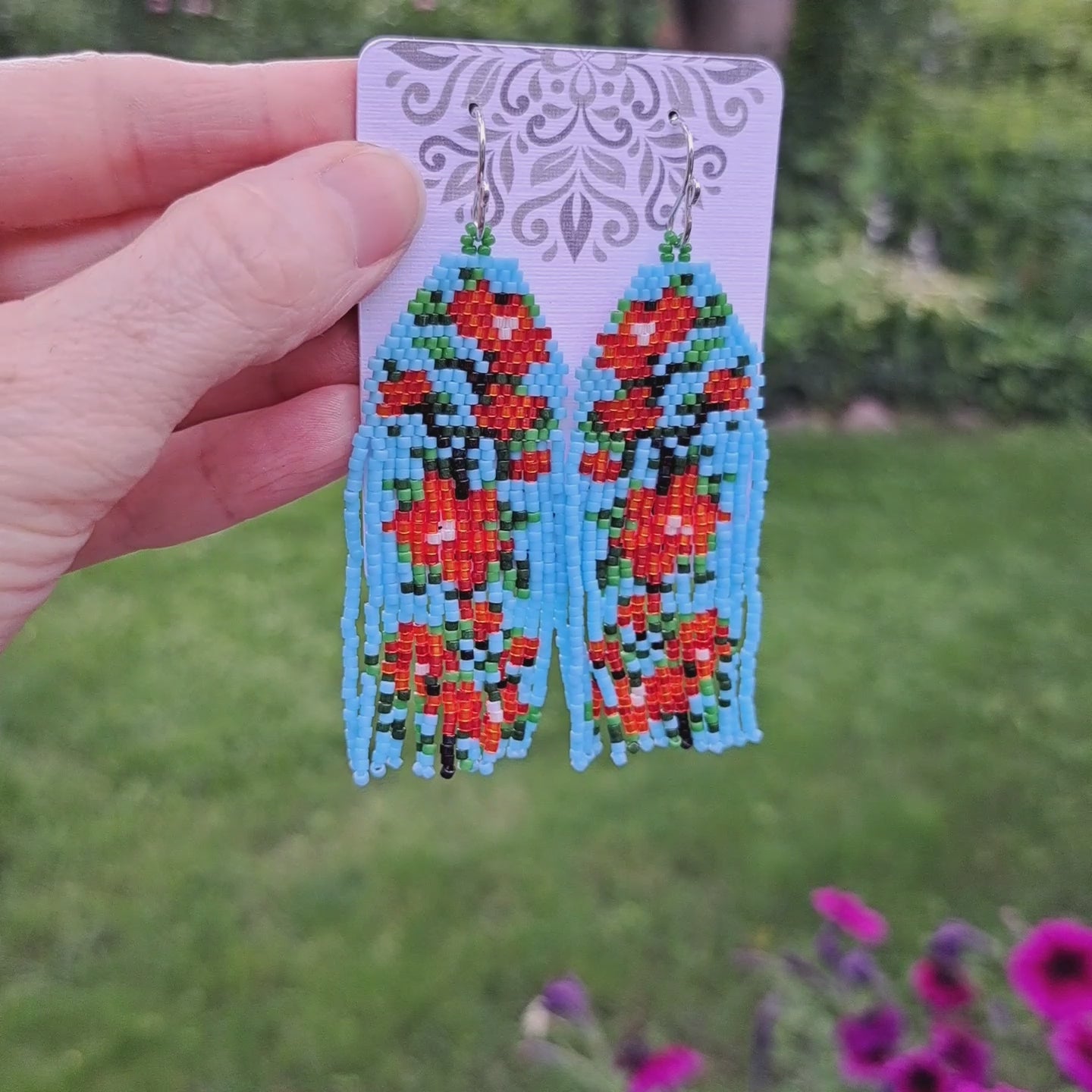 a pair of light blue with red flowers seed bead fringe earrings on card held by hand