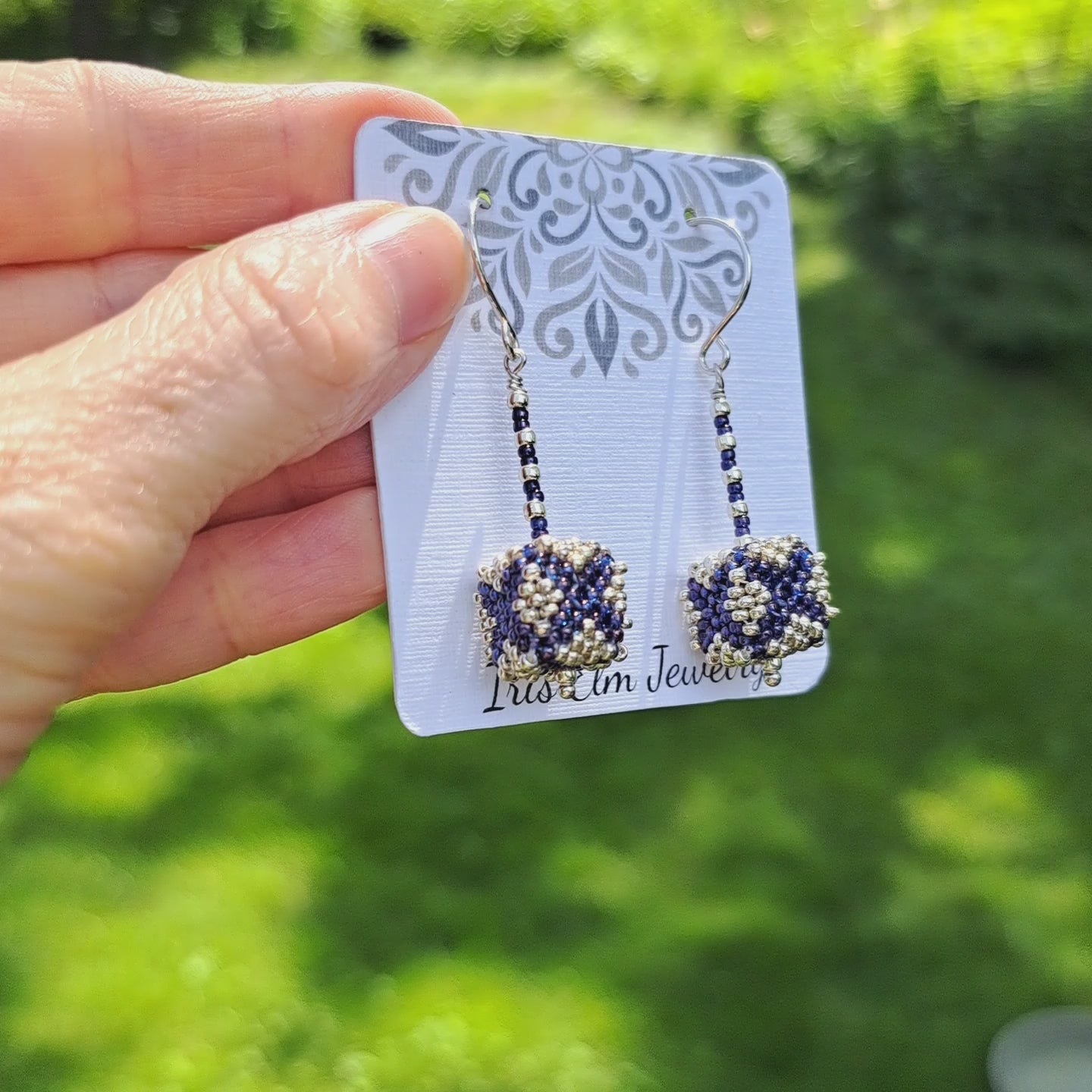 a pair of purple and silver seed bead cube shaped drop earrings on a Iris Elm Jewelry branded card held by hand