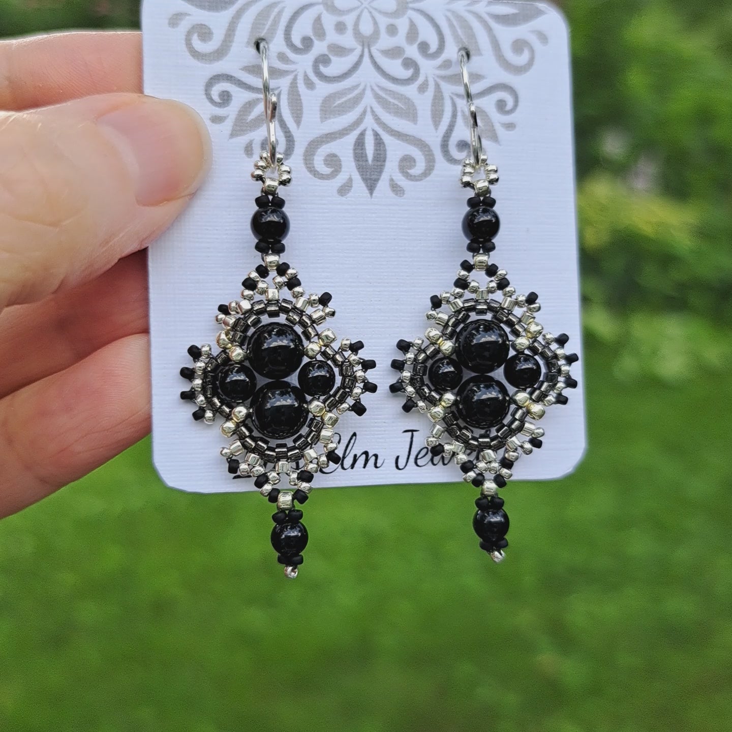 a pair of black onyx beadwork earrings with silver and grey seed bead accent beads on white card held by hand in outdoor setting 