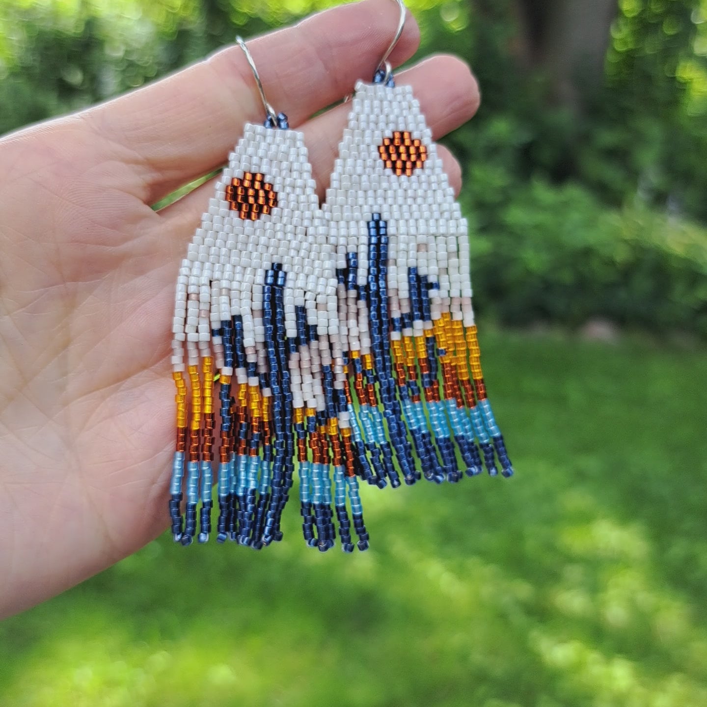 hand holding pair of seed bead fringe earrings with cream, blue, and orange sunset cactus design