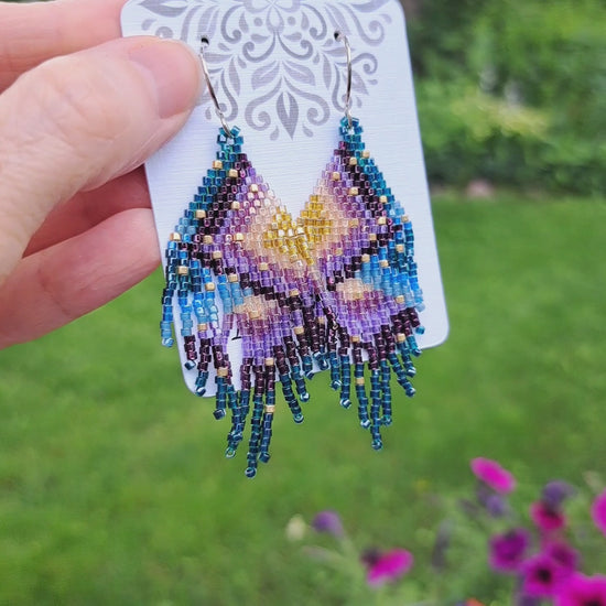 a pair of purple green blue gold seed bead fringe earrings on card held by hand in outdoor setting