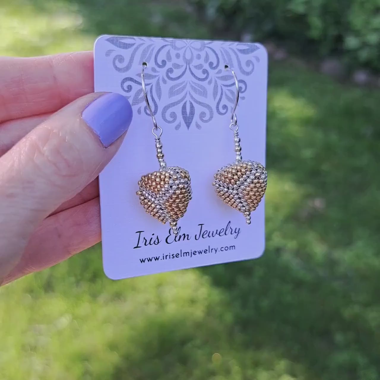 a pair of three dimensional gold and silver seed bead triangle sterling silver earrings on a 'Iris Elm Jewelry' branded card held by hand in outdoor setting