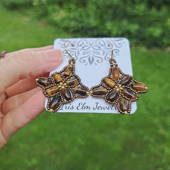 a pair of brown tiger eye beadwork earrings on a 'Iris Elm Jewelry' paper card held by hand in outdoor setting