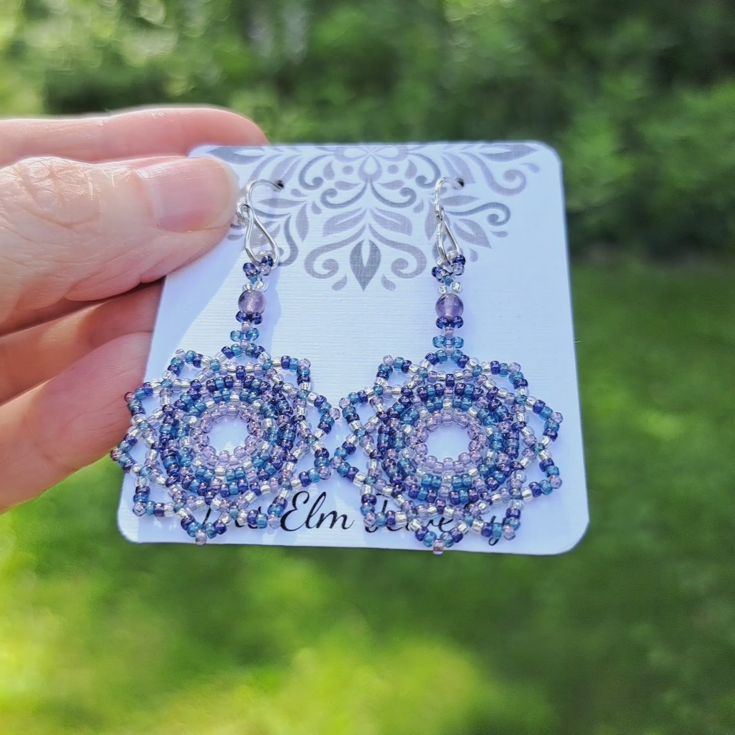 a pair of purple and blue seed bead mandala earrings with amethyst stones on branded Iris Elm Jewelry card held by hand