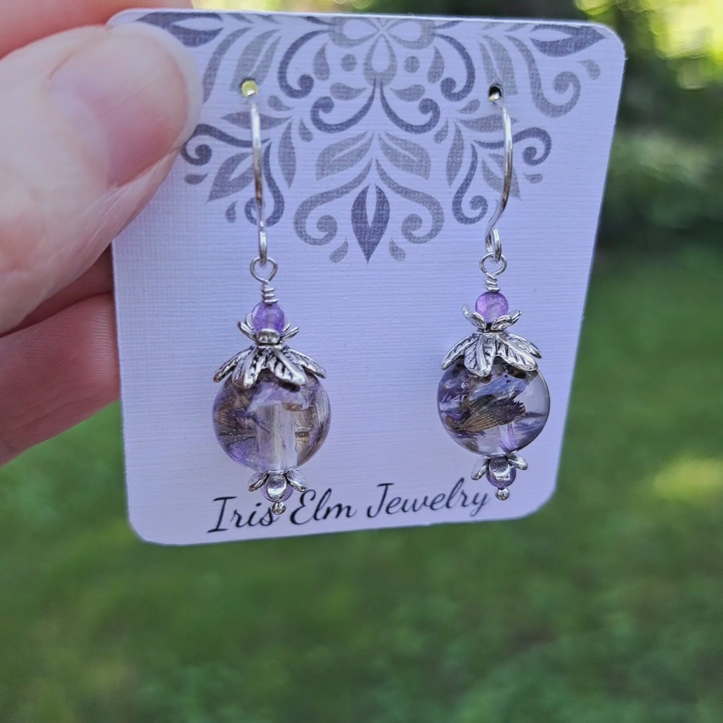 a pair of purple flowers in resin earrings with amethyst stone and silver flower beadcaps. earrings are on earrings card labeled Iris Elm Jewelry held in hand