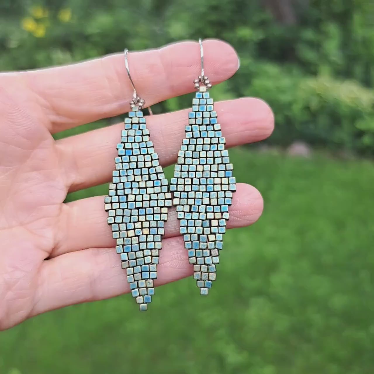 a pair of metallic matte turquoise green glass beadwoven diamond shape earrings held in hand in outdoor setting